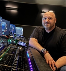 A man with a beard sits at a professional audio mixing desk in a dark studio environment.