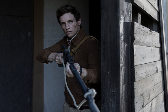 A man in a brown jacket aims a long rifle while peering out from a wooden doorway.