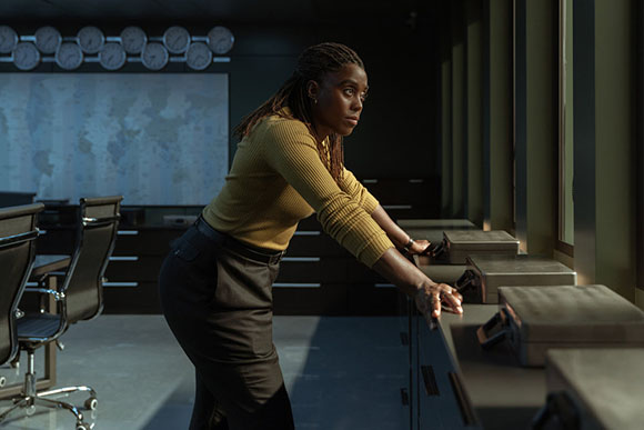 A woman with braided hair leans forward over a desk in a dark, modern office setting.