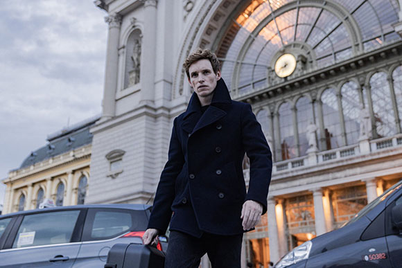 A man in a dark navy pea coat stands before a grand building with a large arched glass window and clock.