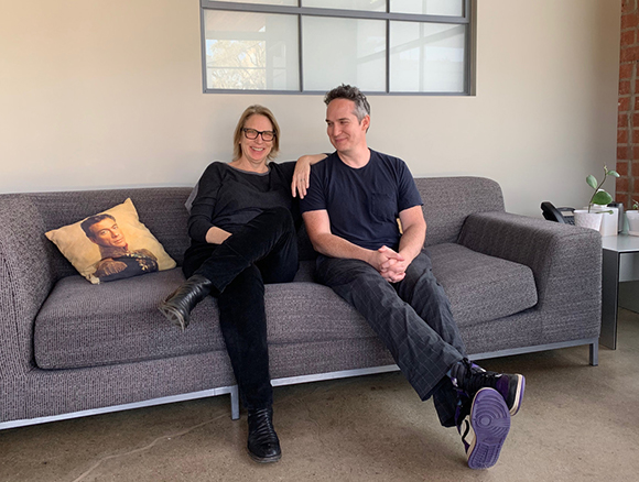 A man and a woman sit together on a grey couch in a bright room.