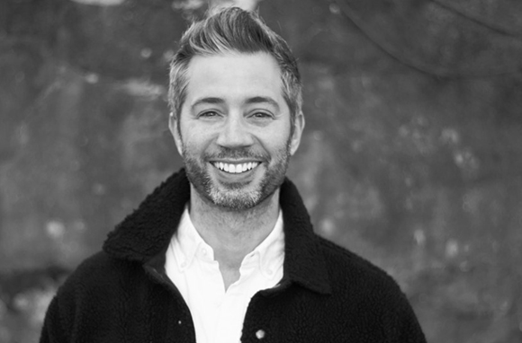 Black and white portrait of a smiling man with short hair and a beard wearing a dark jacket over a white shirt.