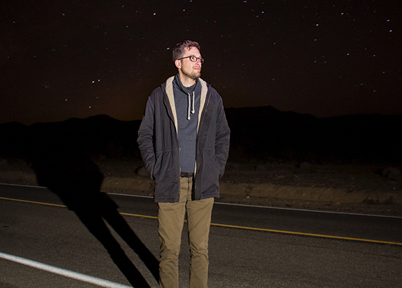 A man in glasses and a jacket stands on a dark road under a starry night sky.