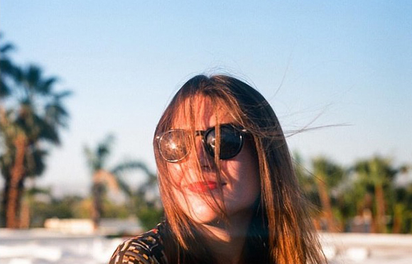 A woman with long brown hair and sunglasses stands outdoors with palm trees in the background.