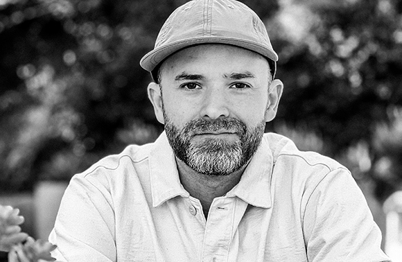 Black and white close-up portrait of a man with a beard wearing a baseball cap and a collared shirt.