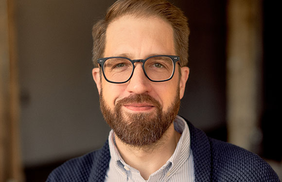 A man with light brown hair, a beard, and black-rimmed glasses looking toward the camera.