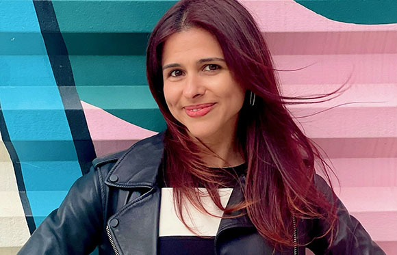 A woman with long reddish-brown hair and a black leather jacket smiles against a pink and teal mural.