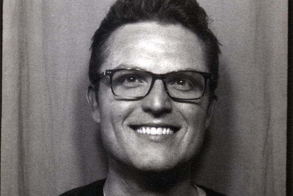 Black and white close-up portrait of a smiling man wearing dark-rimmed glasses.