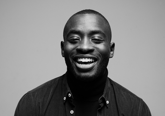 Black and white headshot of a smiling Black man wearing a dark turtleneck and button-down shirt.