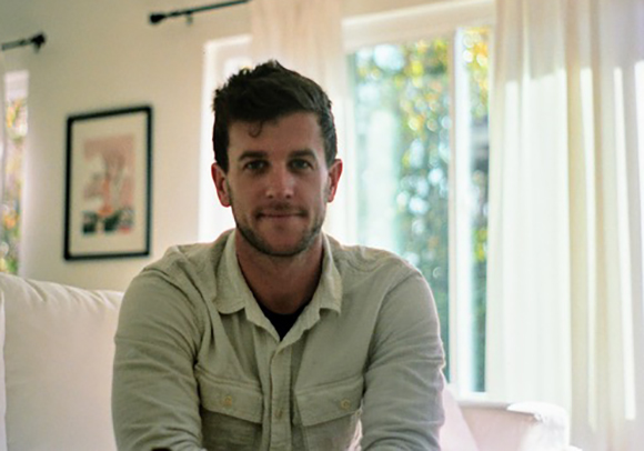 A man with short dark hair and a light-colored button-down shirt sits in a bright room.