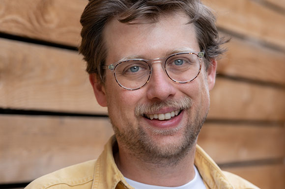 A smiling man with light brown hair and round glasses wearing a yellow shirt against a wooden background.