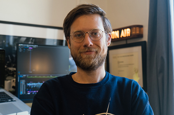 A man with glasses and a beard wearing a dark blue sweatshirt in an office.