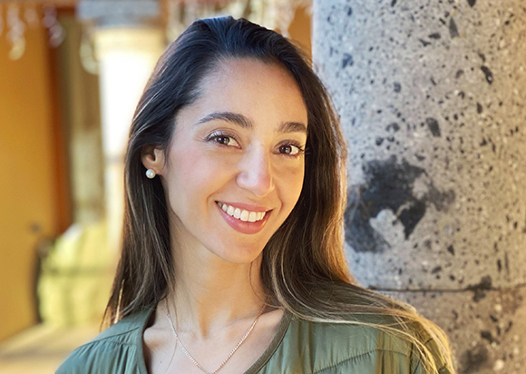 A smiling woman with long dark hair and a green shirt standing next to a stone pillar.