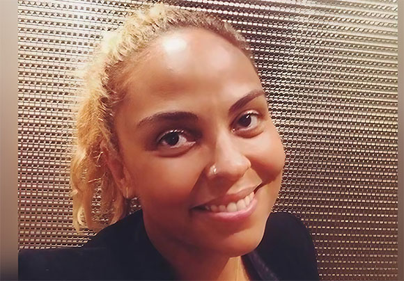 A smiling woman with blonde hair looking toward the camera against a gold textured background.