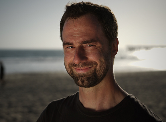 A man with a beard smiling at the camera on a beach during sunset.