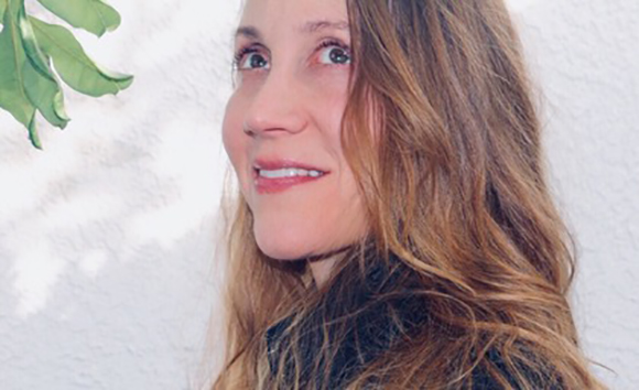A woman with long wavy light brown hair looking back over her shoulder against a white wall.