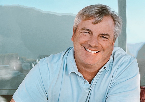 A smiling man with short grey hair wearing a light blue polo shirt against a soft background.