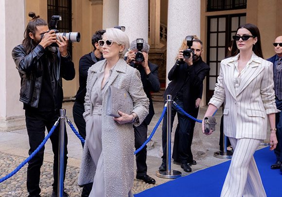 Two women in formal attire walk past several photographers on a blue carpet.