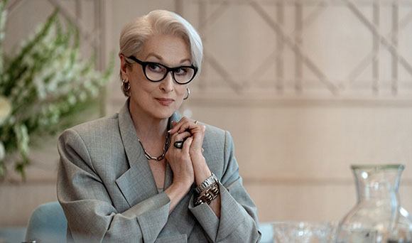 A woman with short white hair and black glasses wearing a grey blazer and jewelry.