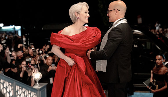 A woman in a voluminous red off-the-shoulder gown smiles at a man in a black suit on a red carpet.