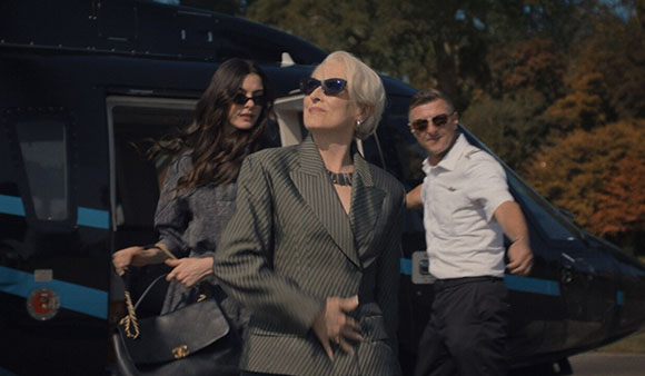 A woman in a grey pinstripe suit stands in front of a helicopter with two people behind her.