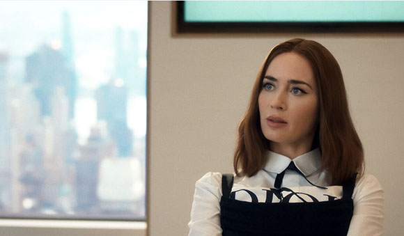 A woman with reddish-brown hair and blue eyes looks upward in an office with a city view.