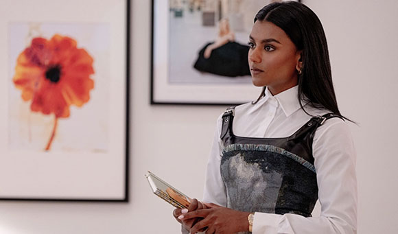 A woman with long dark hair wearing a white collared shirt and a dark vest stands in a room with framed art.