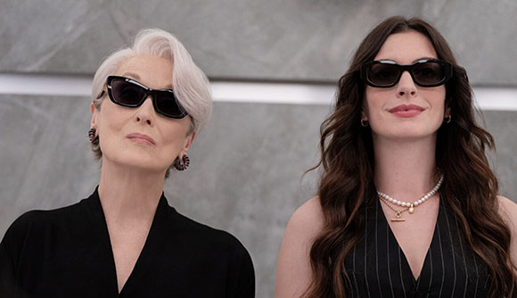 Two women in black outfits and sunglasses standing side by side against a gray background.