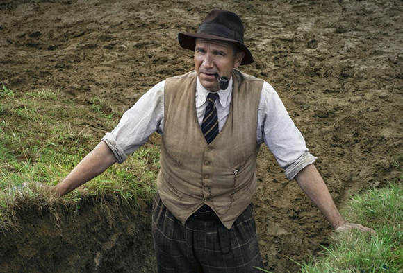 A man wearing a brown hat, tan vest, and tie smokes a pipe while leaning against a dirt embankment.