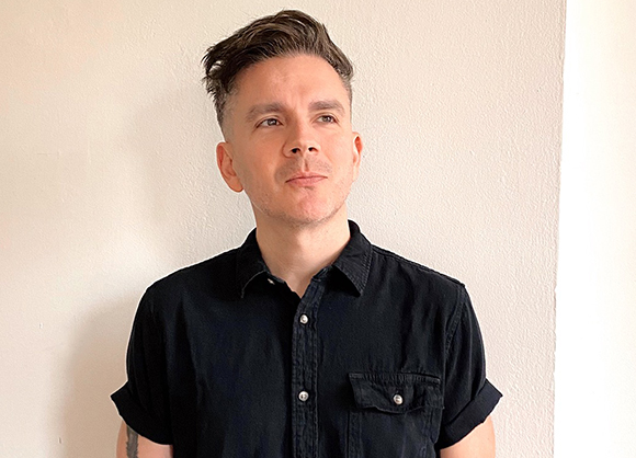 A man with short dark hair wearing a black button-down shirt looking to the side against a plain wall.