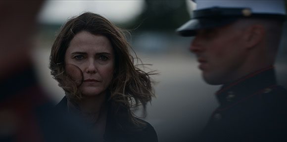 A woman with brown hair looks toward a man in a dark military-style uniform.
