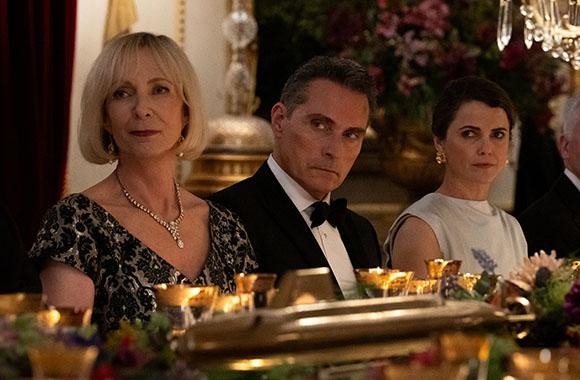 A woman in a black lace dress, a man in a tuxedo, and a woman in a white sleeveless top sit at a formal dinner.