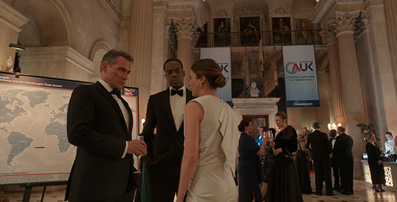 Three people in formal wear talk in a large, ornate hall with high ceilings and marble columns.