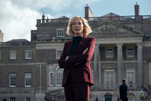 A woman in a maroon suit stands with arms crossed before a large, classical stone building.