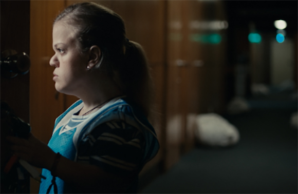 A young person with dark hair in a ponytail looks intently down a dimly lit hallway.