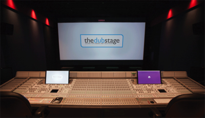 A large audio mixing console in a dark studio with a projection screen displaying 'thedubstage'.