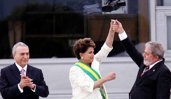 A man and woman hold hands raised in the air while a man in a suit applauds in the background.