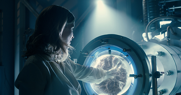 A woman in a lab coat looks into a glowing, circular scientific chamber containing a swirling, textured object.