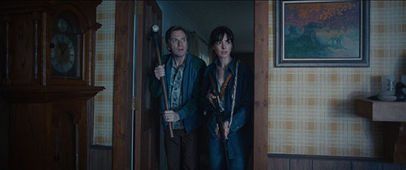 A man and a woman stand in a doorway holding a club and a rifle with worried expressions.