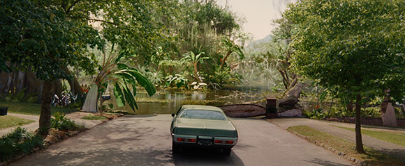 A green vintage car drives down a paved road toward a calm lake surrounded by dense tropical trees and plants.