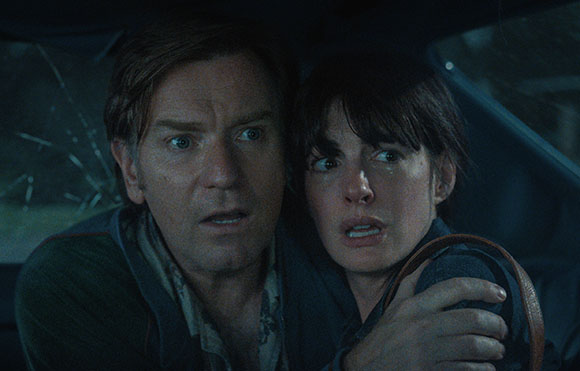 A man and a woman with wide eyes and fearful expressions sit close together inside a dark vehicle.