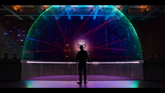 Silhouette of a man at a podium facing a large glowing green sphere and a circular logo with red light rays.