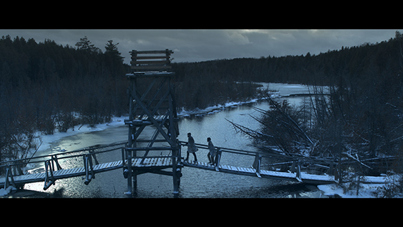 Two people walk across a wooden bridge near a tall wooden tower in a dark, snowy forest landscape.