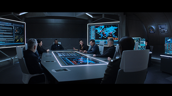 People in dark uniforms sit around a glowing digital table in a futuristic command center with large data screens.