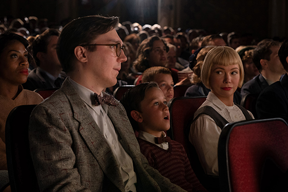 A man in glasses and a bow tie sits among a diverse audience in a dark theater.