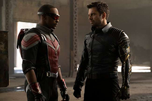 Two men in dark tactical suits stand facing each other in a concrete building.