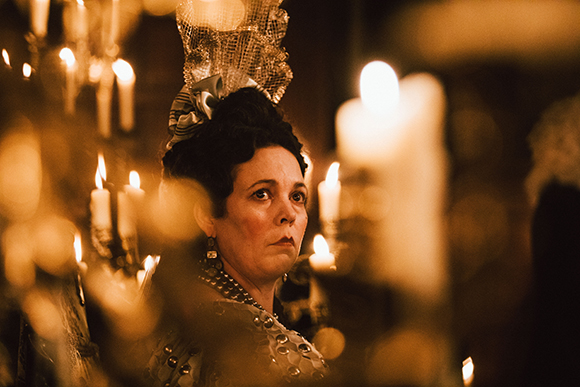 A woman in an ornate historical gown and headpiece looks sideways through blurred candlelight.