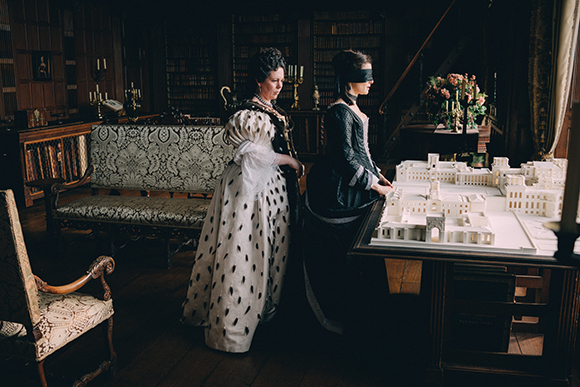 Two women in historical gowns stand in a dark library next to a miniature architectural model.