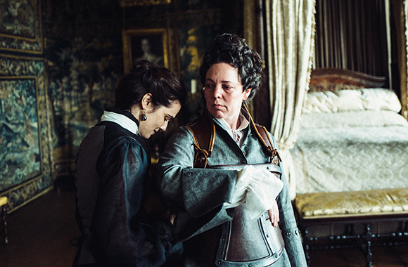 A woman in a dark dress assists another woman into a metal breastplate in a period room.