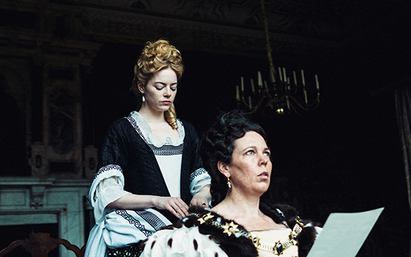A woman in a white and black gown stands behind another woman reading a paper.